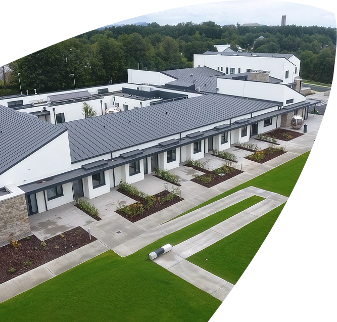 Aerial view of the Mayo Roscommon Hospice, a modern, single-storey white building complex with grey roofs, surrounded by green lawns, trees, and concrete pathways on a cloudy day.