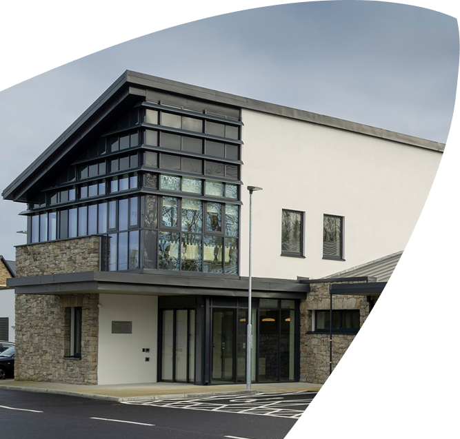 Modern two-storey building with large glass windows, stone accents, and a sloped roof. The paved entrance and parking area in the foreground lead up to the Mayo Roscommon Hospice under a cloudy sky.
