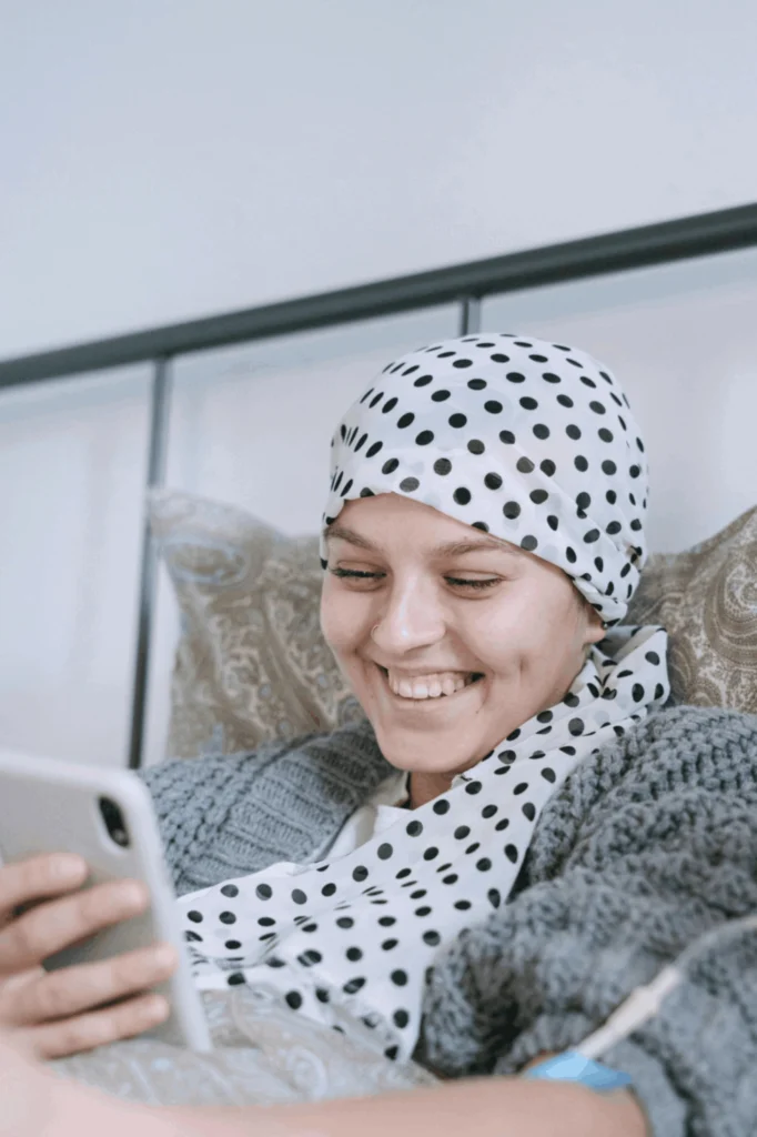 A smiling woman wearing a white and black polka dot headscarf and grey jumper, sits in bed looking at a mobile phone.