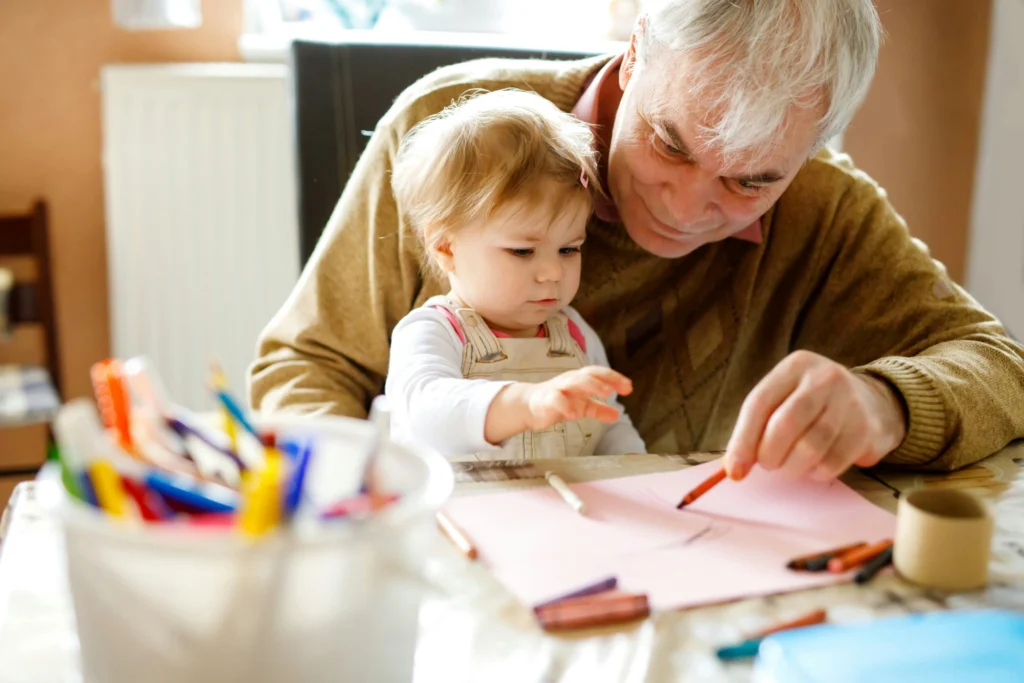 An older man sits with a young child, helping them draw with coloured pencils on pink paper at a table covered in art supplies.