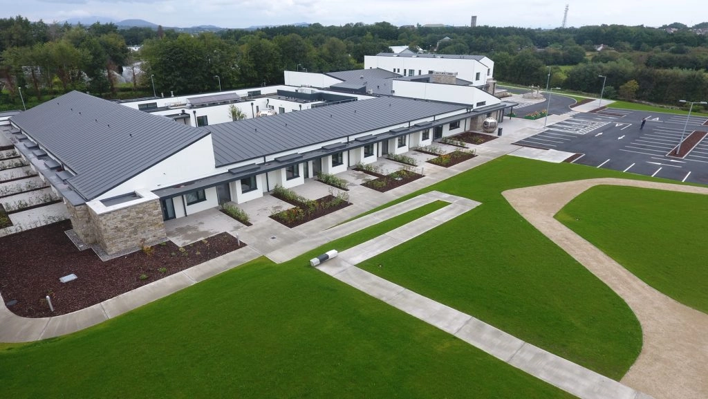 A modern, low-rise building with grey roofs and white walls, Mayo Hospice sits beside tidy green lawns, curved pathways, and a large empty car park, seen from above.