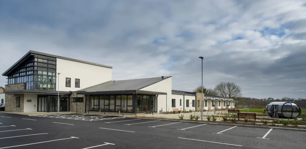 Modern, single-storey building with large windows and stone accents, next to an empty car park and landscaped grounds; this Roscommon Hospice offers compassionate palliative care under a cloudy sky.