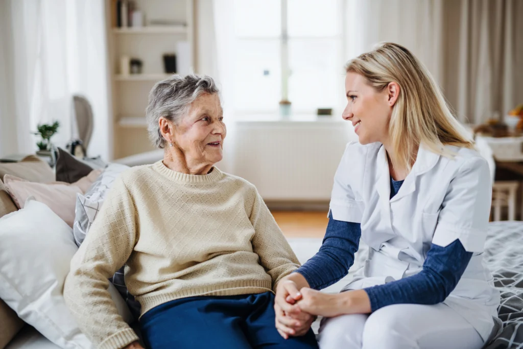 A nurse in white scrubs sits on a sofa holding hands with an elderly woman in a beige jumper, both smiling warmly—capturing the true meaning of palliative care in a bright, cosy living room.