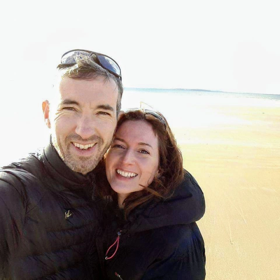 A smiling man and woman pose together on a sandy beach, both wearing black jackets, supporting the Mayo Roscommon Hospice Foundation. The background shows an empty beach and bright sky.