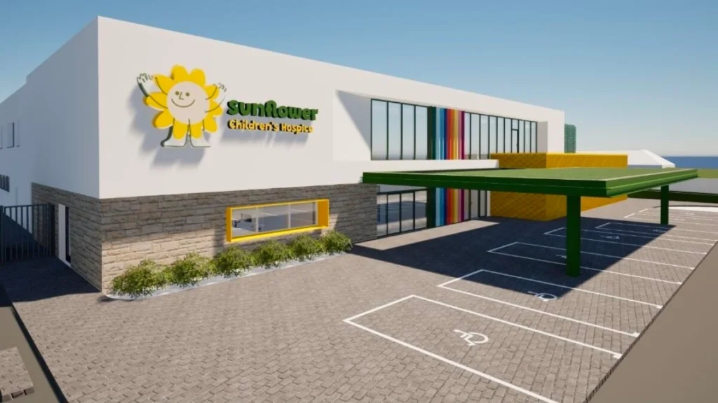 A modern building with a sign reading Sunflower Hospice, a bright sunflower logo, accessible parking spaces, large windows, and colourful vertical accents—providing compassionate hospice care in a welcoming setting.