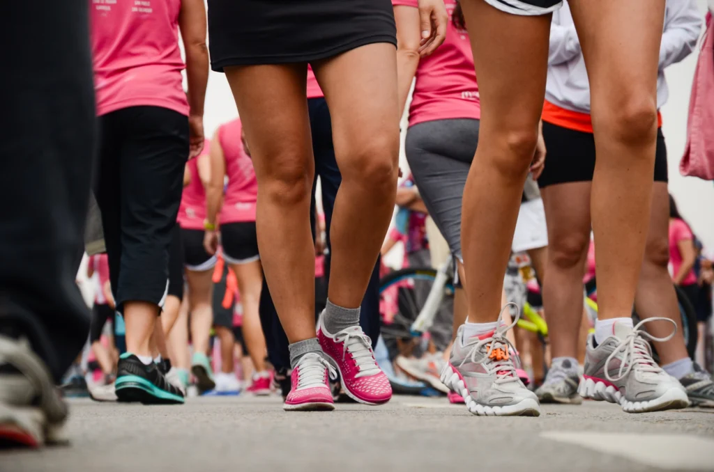 A group of people wearing trainers and pink shirts walk together in an outdoor event, possibly a charity walk or race, with focus on their legs and feet.