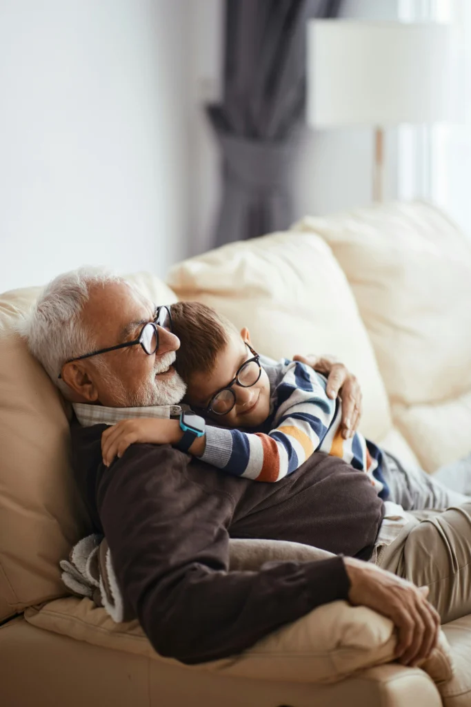 An elderly man and a young boy, both wearing glasses, cuddle on a beige sofa in a warmly lit living room, sharing a heartfelt moment supported by the Mayo Roscommon Hospice Foundation.