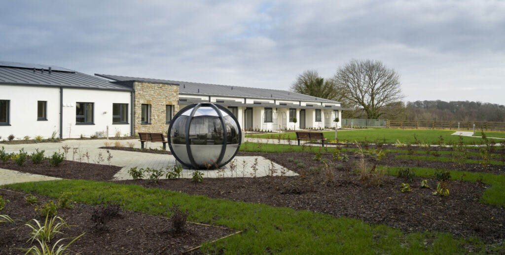 A modern, single-storey building with large windows, a metal roof, landscaped gardens, benches, and a spherical metal sculpture on a cloudy day.