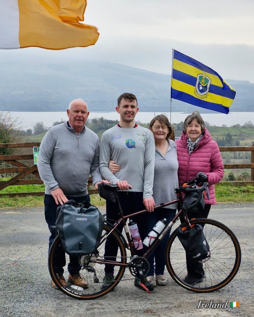 Four people stand outdoors together behind a loaded touring bicycle, showing their support for the Mayo Roscommon Hospice. Two women and two men are smiling, with a lake and mountains in the background. Two flags are flying, including one from County Roscommon, Ireland.