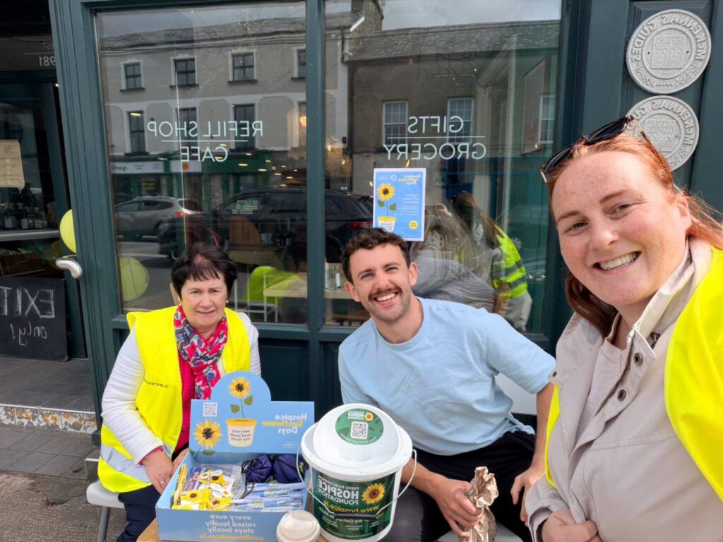 Three people wearing yellow vests smile outside a shop whilst gathering donations for Mayo Roscommon Hospice. They have a charity collection bucket, sunflowers, and donation items on a table. The shop window behind them reads Gifts, Groceries, Refill Shop, Café.