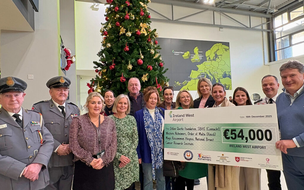 A group of people stand in front of a Christmas tree holding a large cheque for €54,000 from Ireland West Airport to Mayo Roscommon Hospice, surrounded by festive decorations and smiling for the camera.