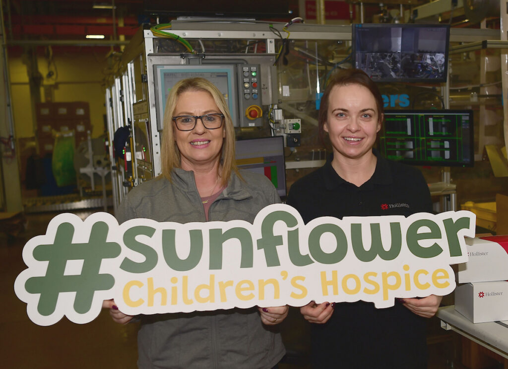 Two women smiling and holding a large #sunflower Children’s Hospice sign inside a room with industrial equipment in the background, showing their support for Mayo Roscommon Hospice.