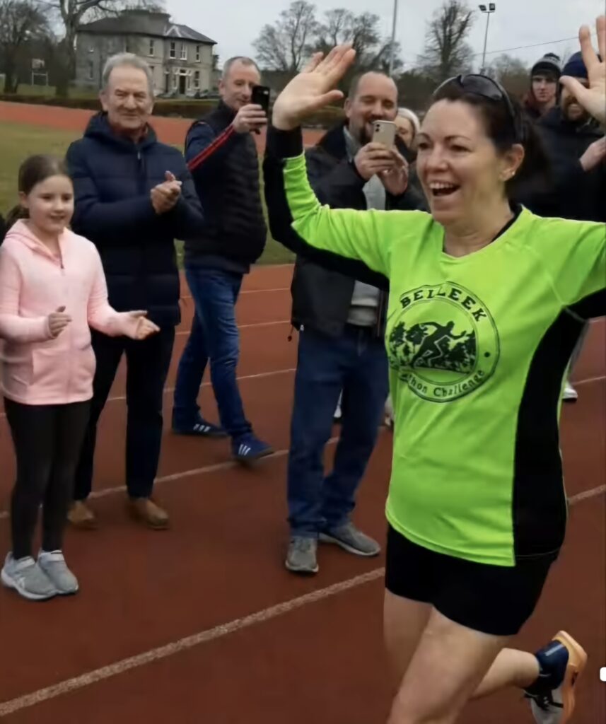 A woman in a bright green shirt finishes a race on a track, smiling with arms raised in triumph, as people on the sidelines clap and cheer in support of Mayo Roscommon Hospice.