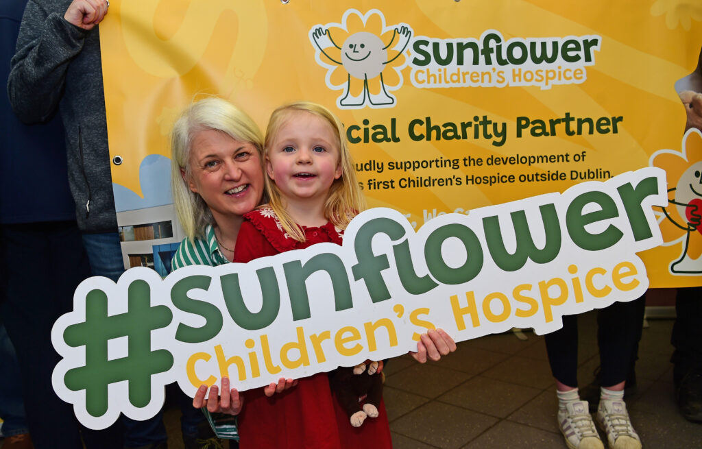 A woman and young girl smile, holding a large Mayo Roscommon Hospice #sunflower Children’s Hospice sign in front of a colourful charity banner.