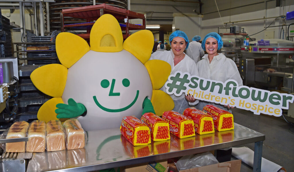 Two women in hairnets and lab coats stand next to a sunflower mascot, holding a #sunflower Children’s Hospice sign in support of Mayo Roscommon Hospice. Packs of bread are on the table in this cheerful bakery setting.