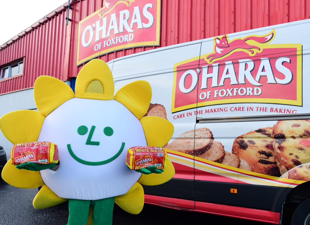 A person in a sunflower costume—symbolic of the Mayo Roscommon Hospice—holds O’Hara’s bread in front of an O’Hara’s of Foxford van and building, both emblazoned with large logos and vibrant images of baked goods.