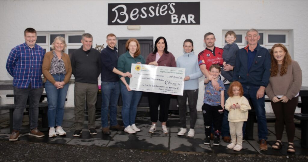 A group of people, including adults and children, stand in front of Bessies Bar holding a large presentation cheque for Mayo Roscommon Hospice. They are smiling and gathered closely together outside the building.