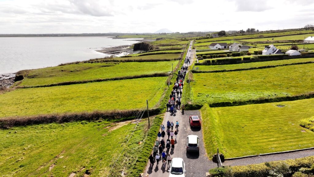A group of people walk along a rural road beside the coast, passing green fields and houses under a cloudy sky. Cars are parked on the roadside as supporters take part in a charity walk for Mayo Roscommon Hospice.