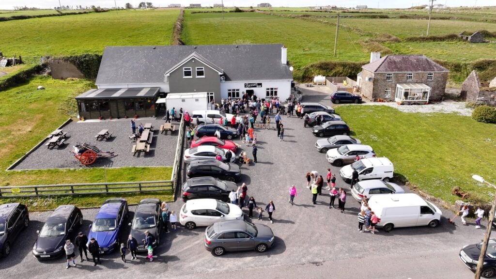 A crowd gathers outside the grey Mayo Roscommon Hospice building with many cars parked around it. People stand in queues and groups, while green fields and a few other buildings are visible in the background.