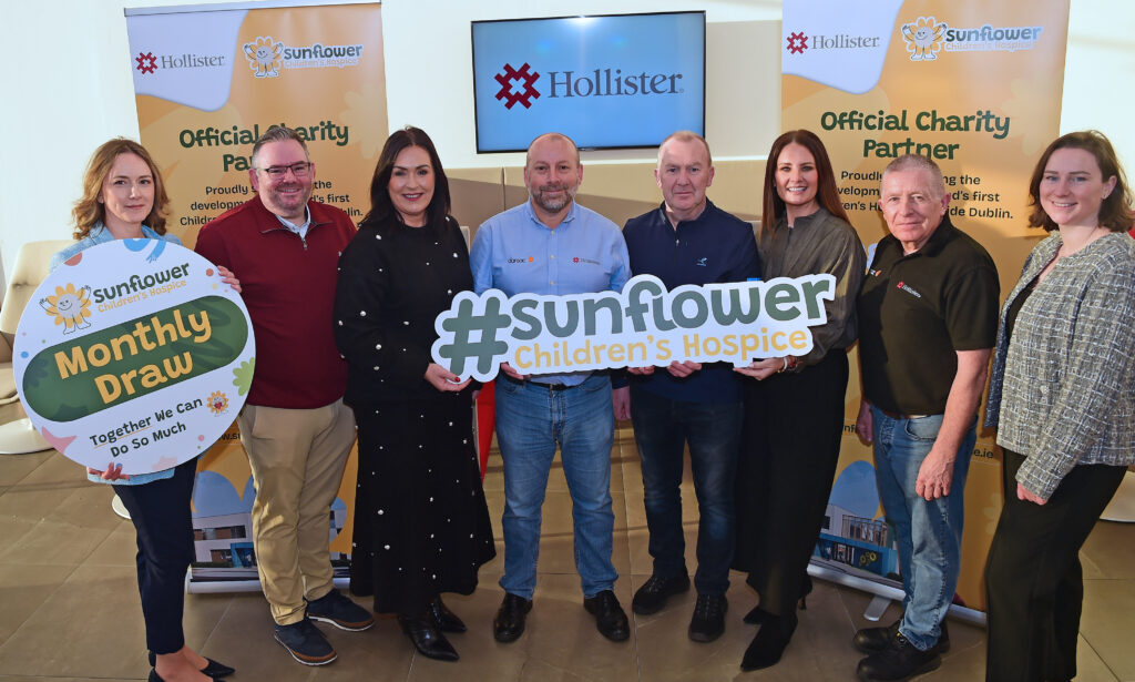 Eight people stand indoors, smiling and holding signs reading #sunflower Children's Hospice and Monthly Draw. Banners for Sunflower Children's Hospice, Mayo Roscommon Hospice, and Hollister are displayed in the background.