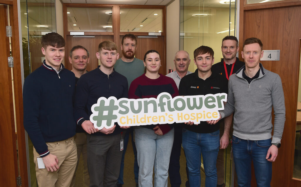 Nine people stand indoors, smiling at the camera. The front row holds a large #sunflower Children’s Hospice sign, proudly supporting Mayo Roscommon Hospice. The background shows glass doors and wooden panelling.