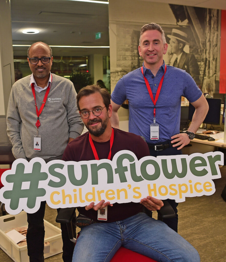 Three men wearing red lanyards smile in an office, with one seated holding a large sign that reads #sunflower Children’s Hospice, highlighting their support for the Mayo Roscommon Hospice.