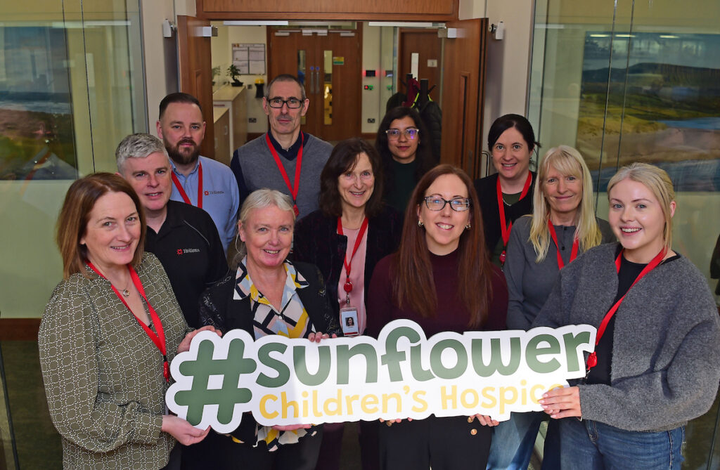 A group of people stand indoors, smiling and holding a large sign that reads #sunflower Children's Hospice. Most are wearing red lanyards, showing their proud connection to Mayo Roscommon Hospice.