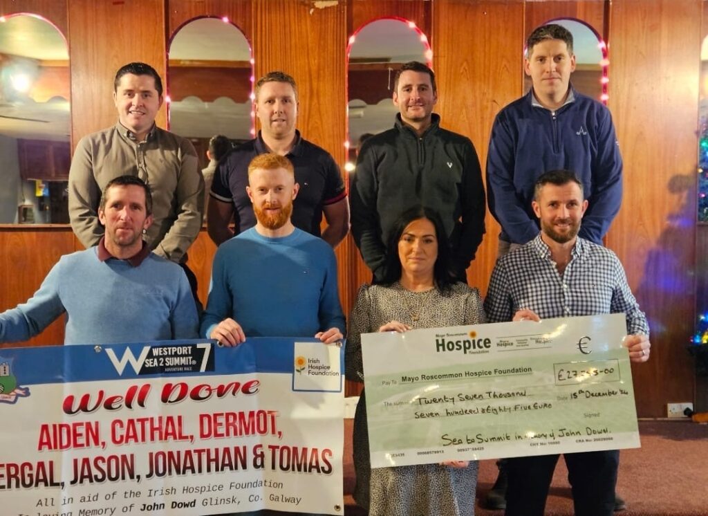 Eight people pose indoors, holding a large banner and an oversized cheque for €2,350 made out to the Mayo Roscommon Hospice Foundation. The mood is celebratory, with fairy lights and wooden walls in the background, as friends and supporters gather to show their enthusiasm for the Mayo Roscommon Hospice’s important work.