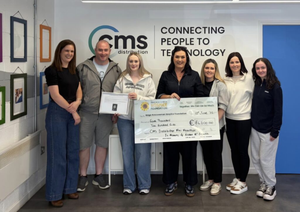 Seven people stand in a row smiling, one holding an oversized donation cheque for €1,600 made out to Mayo Roscommon Hospice and another holding a certificate. A CMS Distribution sign is on the wall behind them.