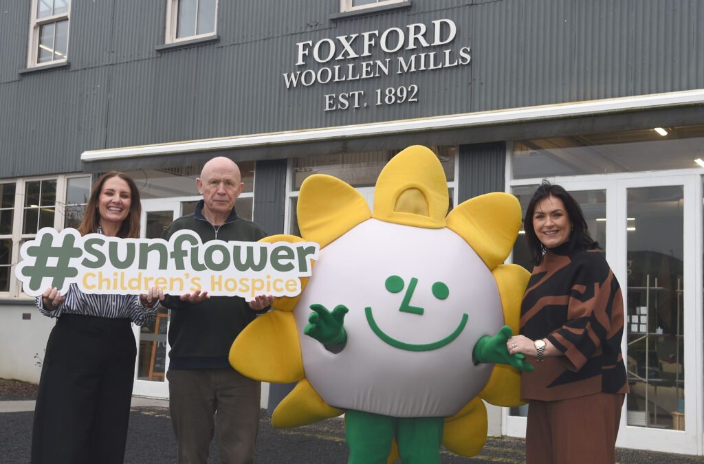 Three adults and a person in a sunflower mascot costume stand smiling in front of Foxford Woollen Mills, holding a #sunflower Children’s Hospice sign, showing their support for Mayo Roscommon Hospice.