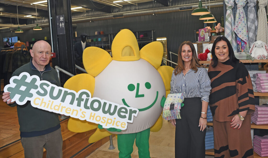 Four people stand in a shop, including one in a sunflower costume. One person holds a large sign reading #sunflower Children’s Hospice, highlighting their support for causes like the Mayo Roscommon Hospice. Blankets and soft toys are visible in the background.