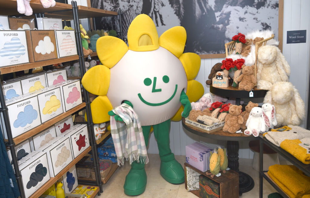 A person in a large, round, yellow and white flower mascot costume stands in a shop holding a scarf, surrounded by shelves of wool, soft toys, and Mayo Roscommon Hospice gift items.