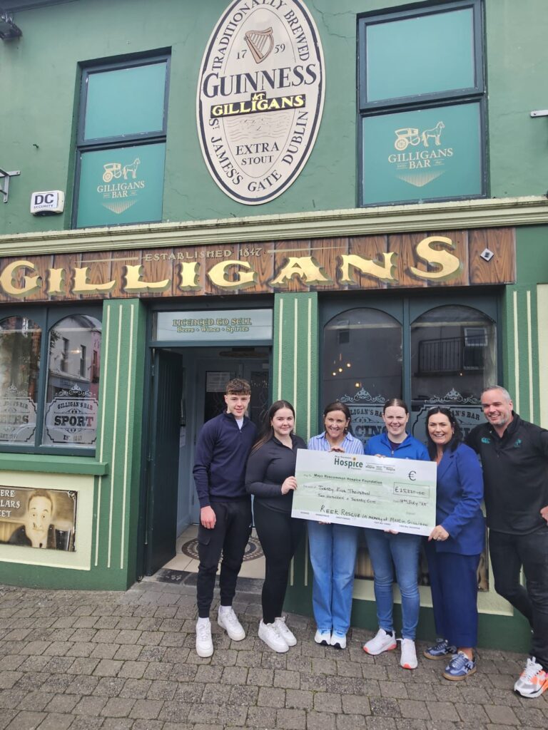 Six people stand outside Gilligans Bar holding a large donation cheque for the Mayo Roscommon Hospice. The bar has green walls and prominent Guinness signage above the entrance. Everyone is smiling at the camera.