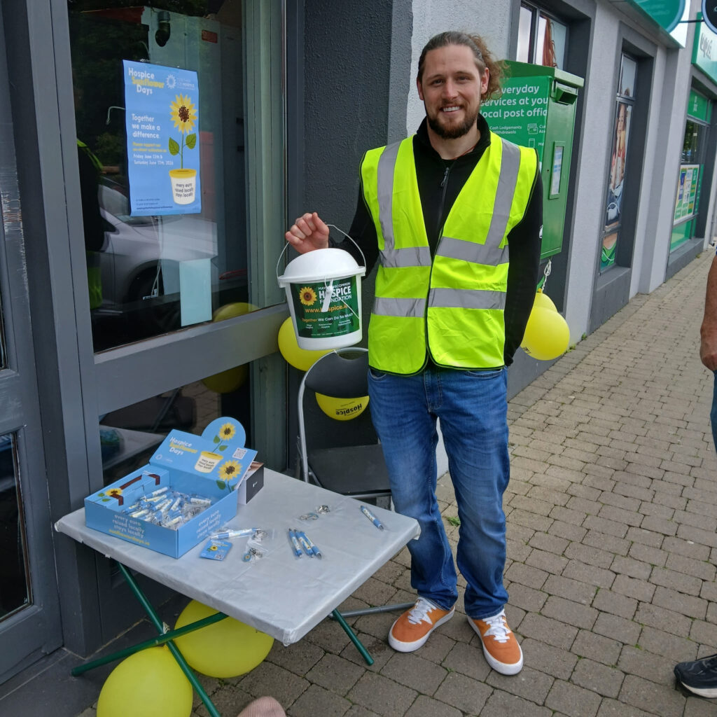 A man in a yellow safety vest holds a Mayo Roscommon Hospice charity bucket and stands by a table with pens and badges outside a building decorated with yellow balloons.